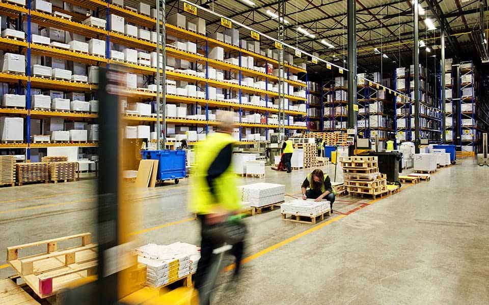 Großes Lager mit hohen Regalen voller Waren, in dem Mitarbeiter in Warnwesten mit der Kommissionierung und Verpackung von Produkten beschäftigt sind.