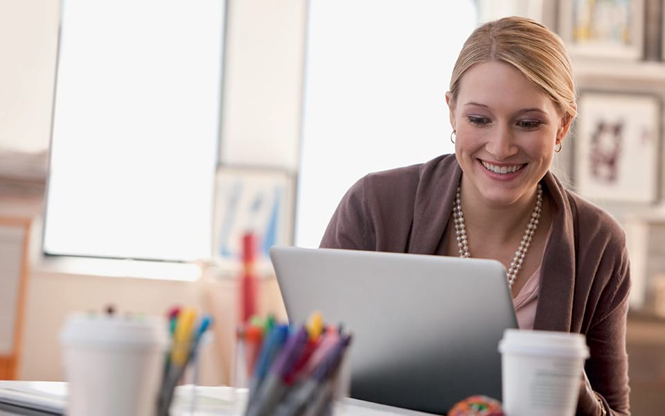 Lächelnde Frau arbeitet an einem Laptop in einem modernen Büro, mit Kaffeebecher und Stiften auf dem Schreibtisch und nutzt Antalis elektronische Bestellmöglichkeiten